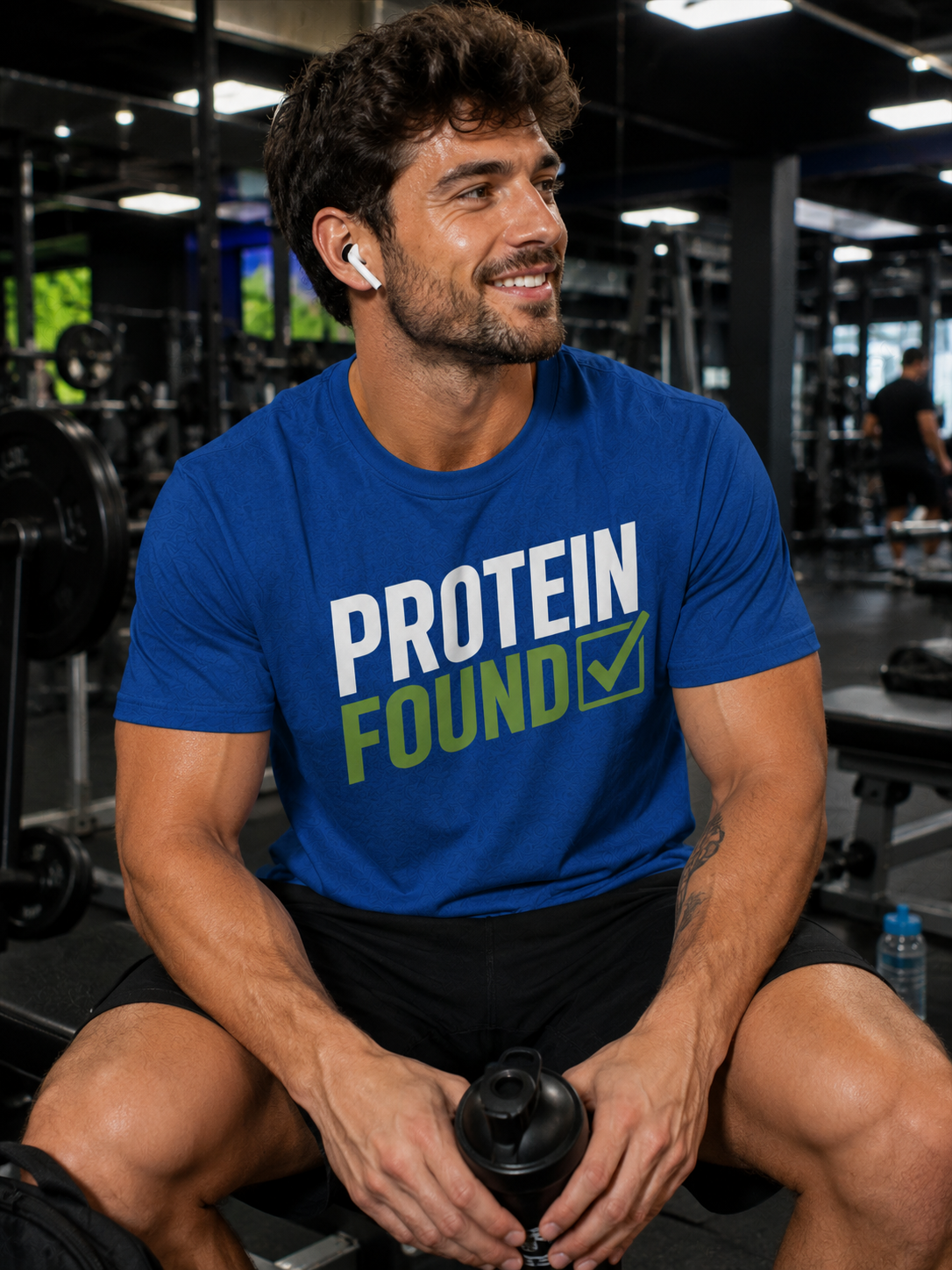 A man in a gym wearing a blue t-shirt with the text 'PROTEIN FOUND' in white and green, holding a black shaker bottle.