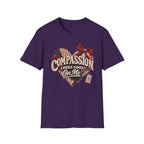 A purple short-sleeve crewneck t-shirt with a graphic on the front. The graphic features a heart made of plaid and flag patterns, a red bow, and the text 'COMPASSION LOOKS GOOD On Me' in a vintage style.