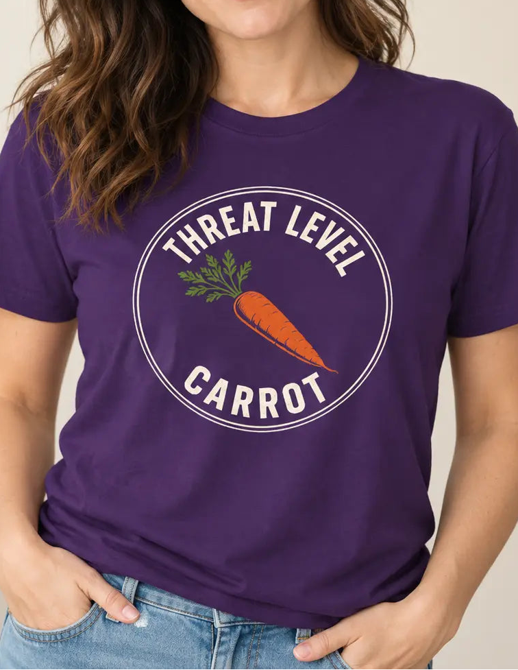 A woman wearing a purple short-sleeve graphic t-shirt with a circular 'Threat Level Carrot' design featuring an orange carrot, paired with blue jeans in a brightly lit indoor setting.