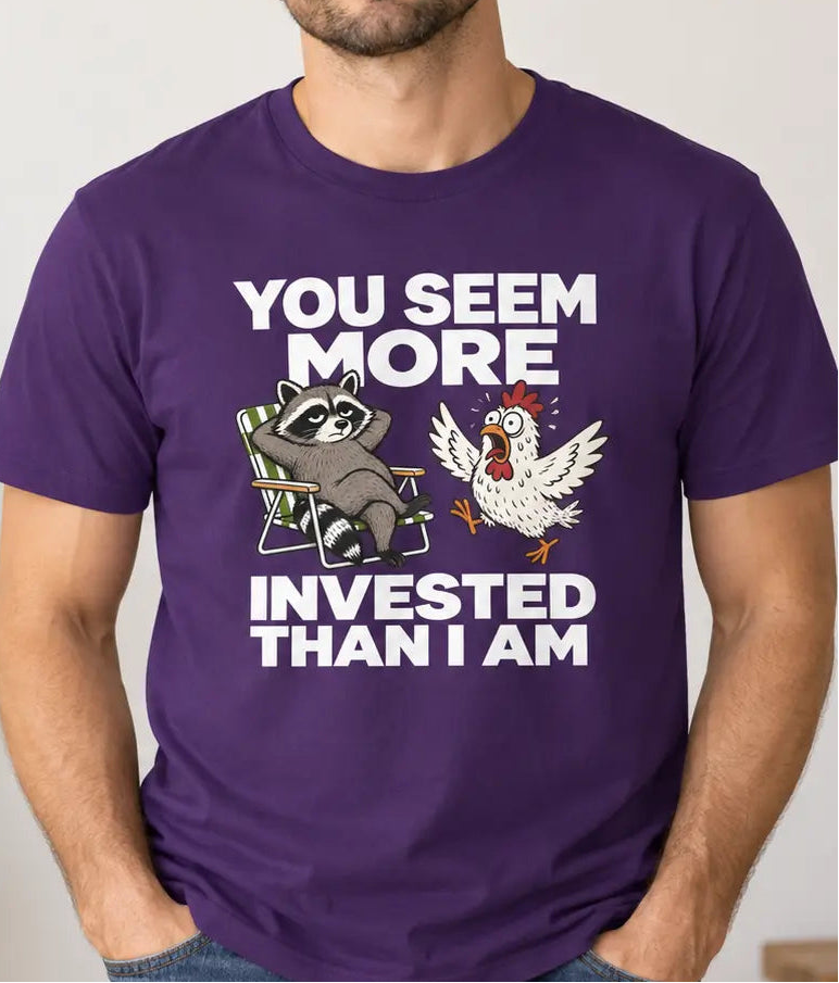 A man wearing a purple graphic t-shirt with a raccoon and chicken illustration and the text 'YOU SEEM MORE INVESTED THAN I AM'.