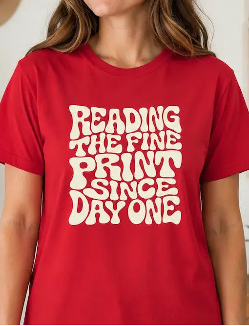 A woman wearing a red graphic t-shirt with the text 'READING THE FINE PRINT SINCE DAY ONE' in a retro white font, standing in a cozy, boho-style room with plants and a macrame wall hanging.