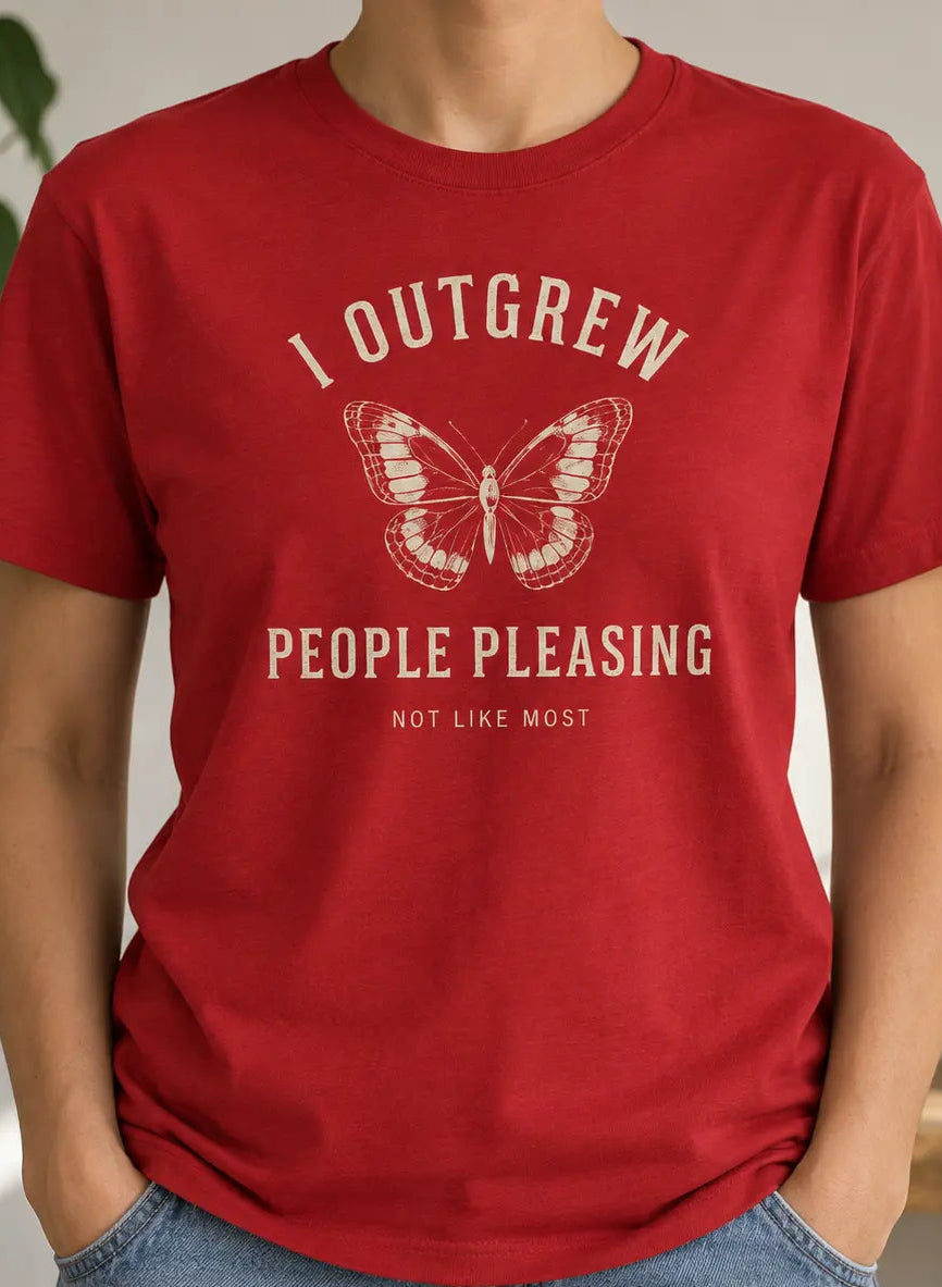 A person wearing a red short-sleeve graphic T-shirt with a butterfly and the text 'I OUTFREW PEOPLE PLEASEING NOT LIKE MOST' in a light cream print, paired with blue jeans in a softly lit indoor setting with plants and a shelf in the background.