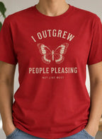 A person wearing a red short-sleeve graphic T-shirt with a butterfly and the text 'I OUTFREW PEOPLE PLEASEING NOT LIKE MOST' in a light cream print, paired with blue jeans in a softly lit indoor setting with plants and a shelf in the background.
