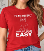 A woman wearing a red graphic t-shirt with a white line art cat wearing glasses and the text 'I'M NOT DIFFICULT YOU'RE JUST USED TO EASY'.