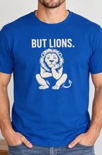 A man wearing a royal blue crewneck t-shirt with a white graphic of a thinking lion and the text 'BUT LIONS.' printed on the chest, standing in a brightly lit room with a plant in the background.