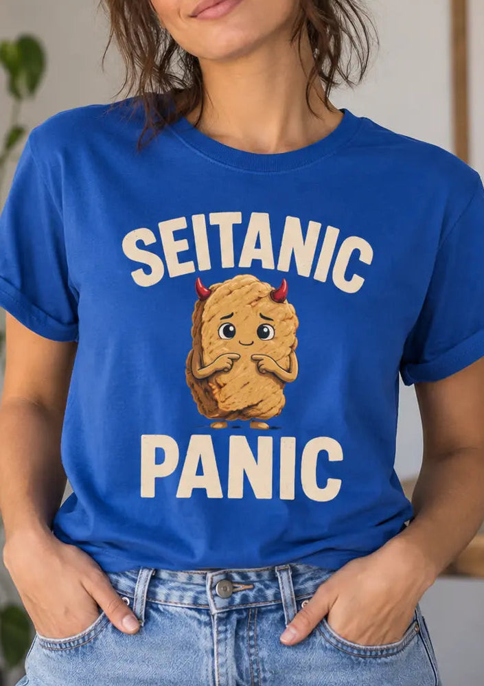 A woman wearing a royal blue graphic t-shirt with the text 'SEITANIC PANIC' and a cartoon character, paired with light wash blue jeans in a cozy indoor setting.