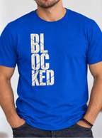 A man wearing a bright blue crewneck t-shirt with the word 'BLOCKED' printed vertically in distressed white block letters on the front, set against a blurred indoor background.