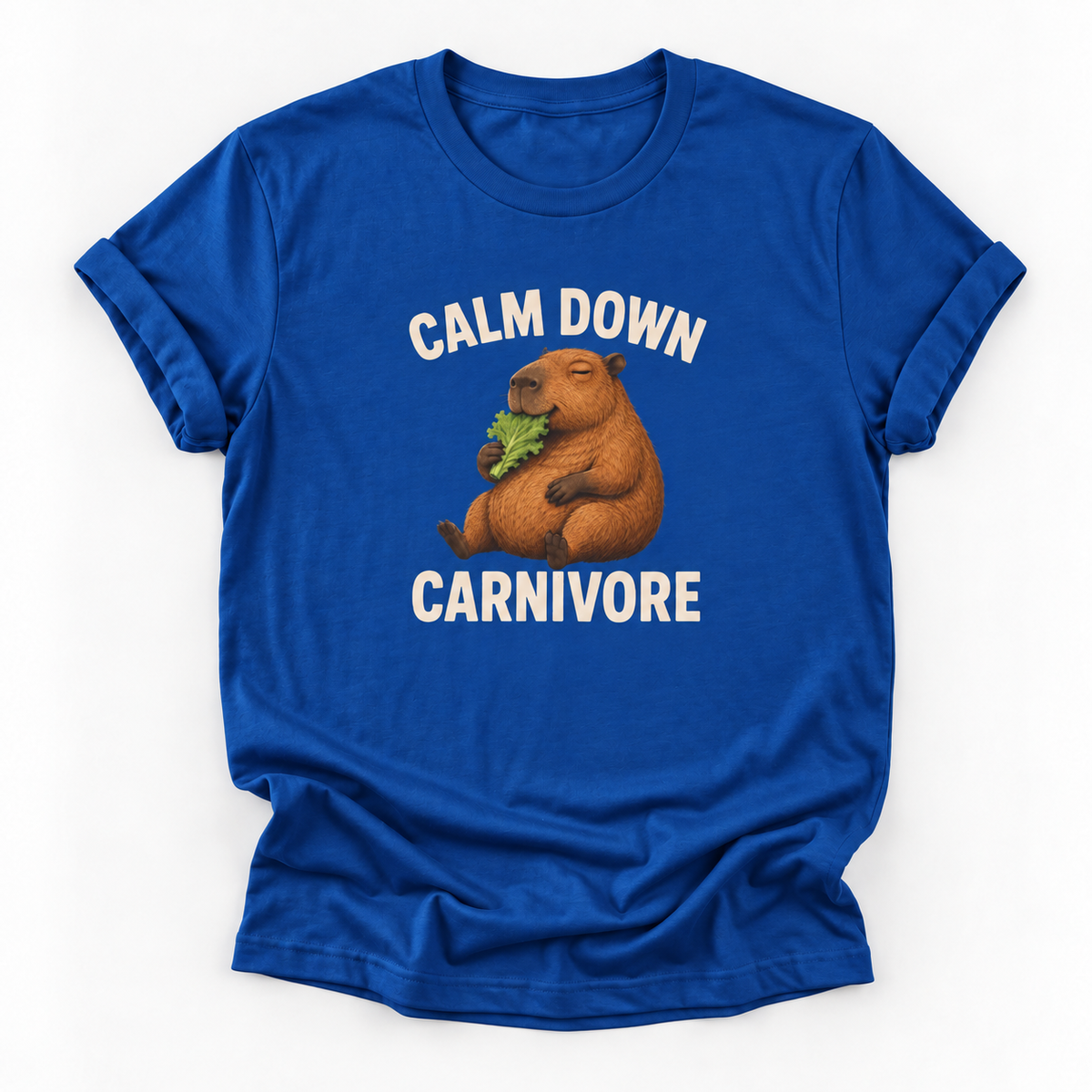 A royal blue short-sleeve t-shirt with a graphic of a capybara eating green leaves, surrounded by the text 'CALM DOWN CARNIVORE' in white. The t-shirt is displayed flat against a clean white background.