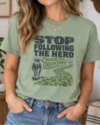 A woman wearing a sage green graphic t-shirt with the text 'STOP FOLLOWING THE HERD CHOOSE THE GREEN PATH' and a cow illustration, posed in a cozy, decorated room.