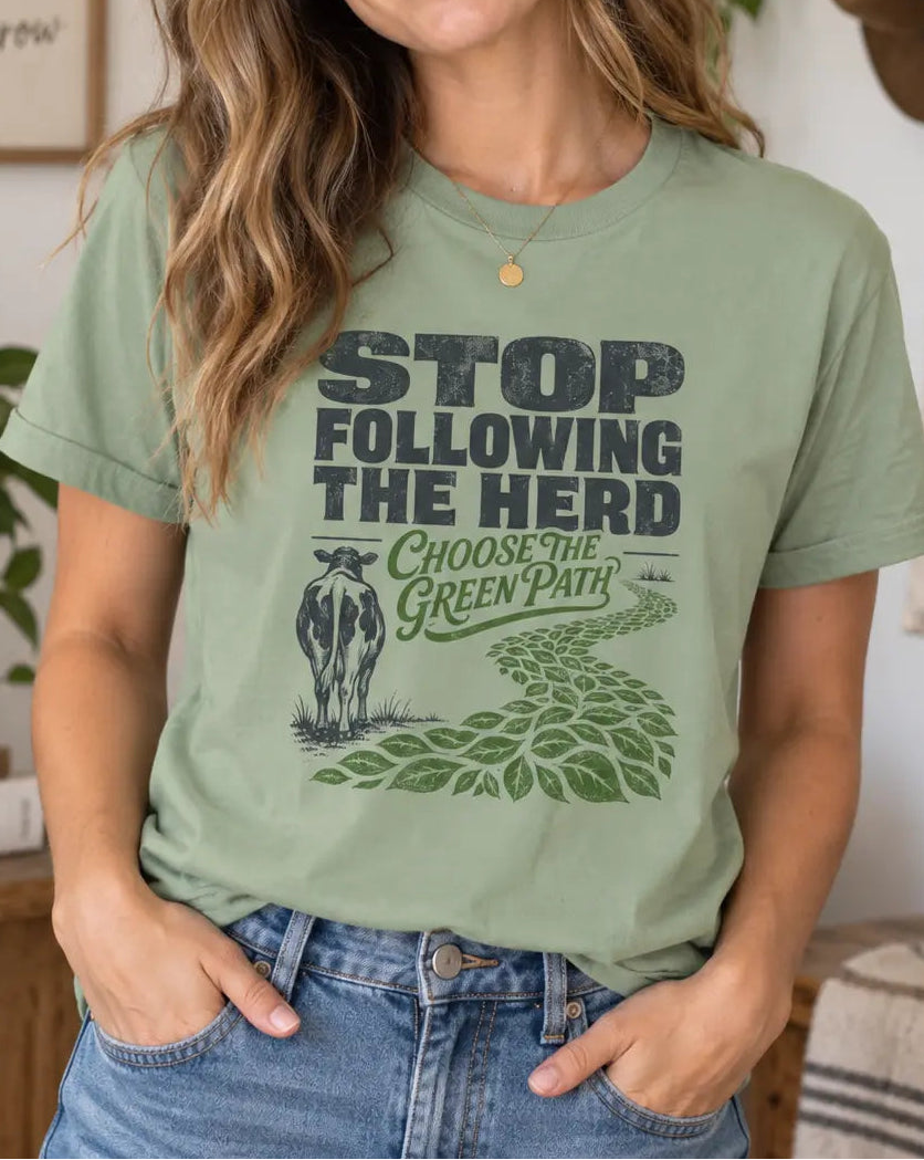 A woman wearing a sage green graphic t-shirt with the text 'STOP FOLLOWING THE HERD CHOOSE THE GREEN PATH' and a cow illustration, posed in a cozy, decorated room.