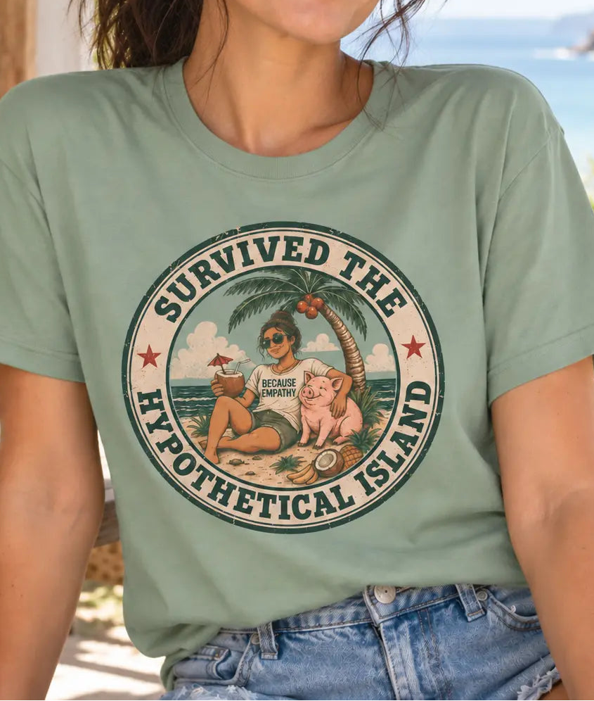 A woman wearing a sage green graphic t-shirt with a circular retro design that reads 'SURVIVED THE HYPOTHETICAL ISLAND' and shows a beach scene with a coconut, sunglasses, and a pink pig. She is sitting outdoors on a patio with a tropical ocean background.