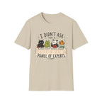 A beige short-sleeve crewneck t-shirt featuring a cartoon graphic of four animals (a cat, frog, panda, and turtle) sitting at a table with books, captioned with the text 'I DIDN'T ASK FOR A PANEL OF EXPERTS.'.