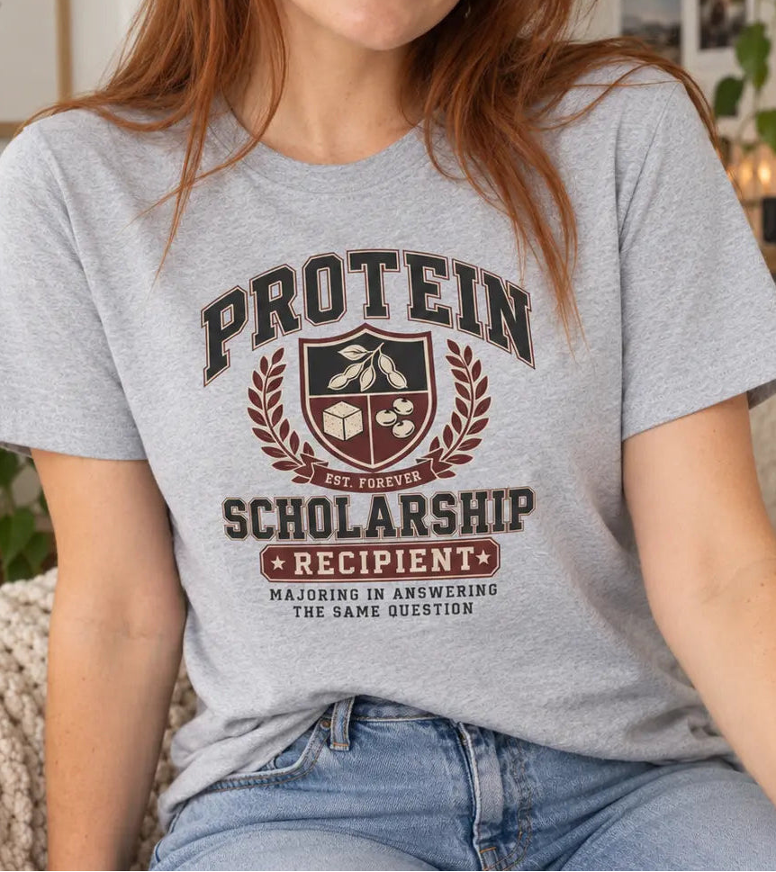 A woman with red hair wearing a light heather gray graphic t-shirt that reads 'PROTEIN SCHOLARSHIP RECIPIENT' with a shield emblem, sitting on a white couch in a cozy room.