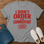 Gray t-shirt with red text 'I Didn't Order the Commentary' on a wooden surface with jeans and sneakers.