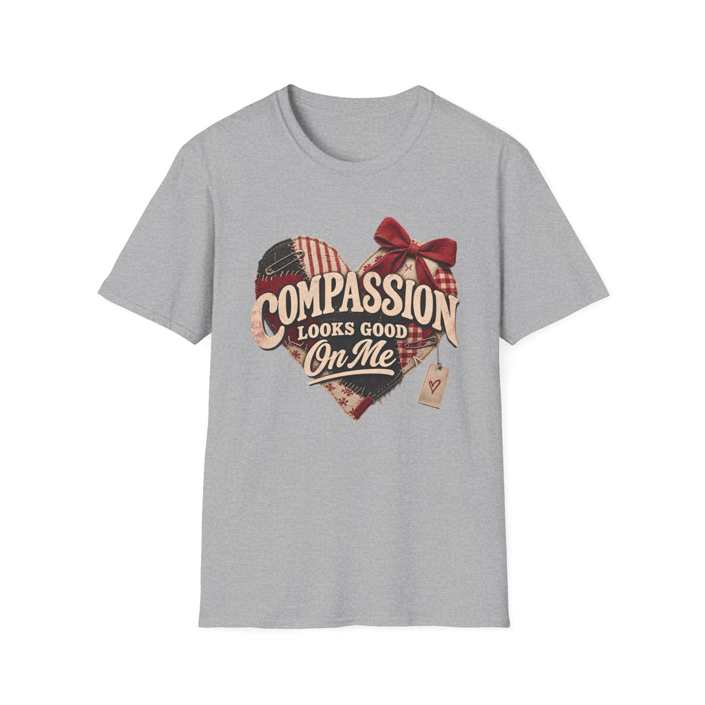 A light gray heather crewneck t-shirt featuring a heart-shaped graphic with the text 'COMPASSION LOOKS GOOD ON ME' in beige script over a red, white, and blue plaid heart design with a red bow and a small hanging tag.