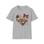 A light gray heather crewneck t-shirt featuring a heart-shaped graphic with the text 'COMPASSION LOOKS GOOD ON ME' in beige script over a red, white, and blue plaid heart design with a red bow and a small hanging tag.