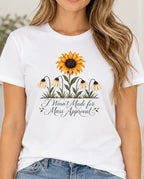 Woman wearing a white graphic T-shirt featuring a bright sunflower above four drooping cream flowers with the words “I Wasn’t Made for Mass Approval” in cursive text.