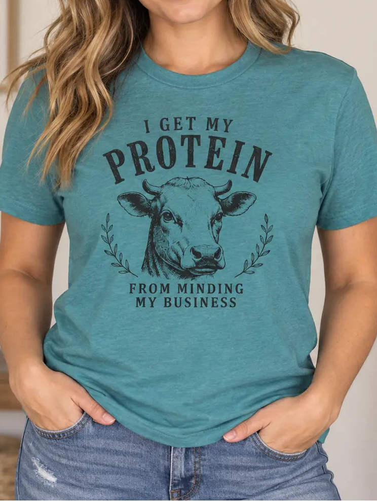A woman wearing a heathered teal graphic t-shirt with a cow illustration and the text 'I GET MY PROTEIN FROM MINDING MY BUSINESS'.
