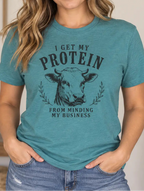 A woman wearing a heathered teal graphic t-shirt with a cow illustration and the text 'I GET MY PROTEIN FROM MINDING MY BUSINESS'.