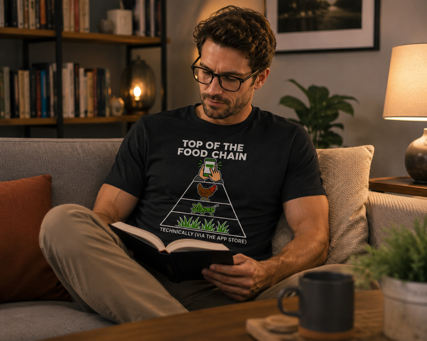 A man wearing glasses sits on a couch in a cozy living room, reading a book. He is wearing a black graphic t-shirt with a white text design that reads 'TOP OF THE FOOD CHAIN' above a pyramid graphic and 'TECHNICALLY (EXA THE APP STORE!)' below.