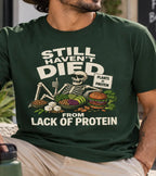 A man wearing a vegan dark green graphic t-shirt with a cartoon skeleton and food illustration that reads 'STILL HAVEN'T DIED FROM LACK OF PROTEIN'.