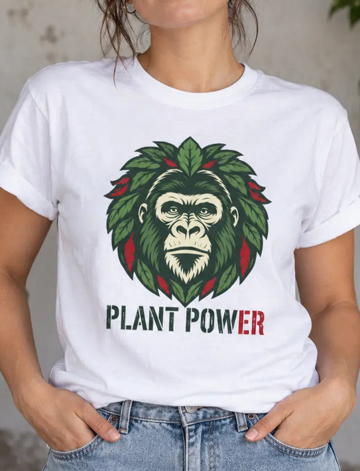 A woman wearing a white graphic t-shirt with a green gorilla face surrounded by leaves and the words 'PLANT POWER' in green and red, paired with blue jeans, standing in front of a rustic wall with plants.