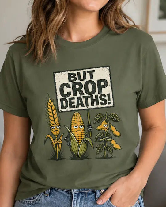A woman wearing an olive green graphic t-shirt with a cartoon corn design and the text 'BUT CROP DEATHS!' in a cozy living room setting.