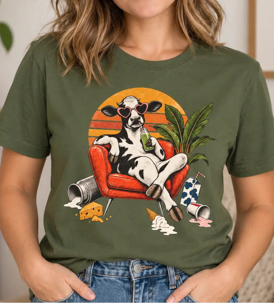 A woman wearing an olive green graphic t-shirt with a cow lounging in an armchair design, posed in a cozy indoor setting.