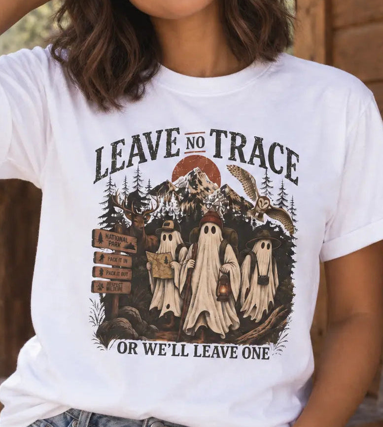 A woman wearing a white graphic t-shirt with a Halloween-themed ghost design and the text 'LEAVE NO TRACE OR WE'LL LEAVE ONE', paired with blue jeans and a brown beanie, standing on a rustic wooden porch.