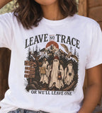 A woman wearing a white graphic t-shirt with a Halloween-themed ghost design and the text 'LEAVE NO TRACE OR WE'LL LEAVE ONE', paired with blue jeans and a brown beanie, standing on a rustic wooden porch.