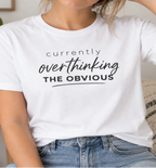 A woman wearing a white graphic t-shirt with the text 'currently overthinking THE OBVIOUS' in black script, paired with blue jeans in a cozy indoor setting.