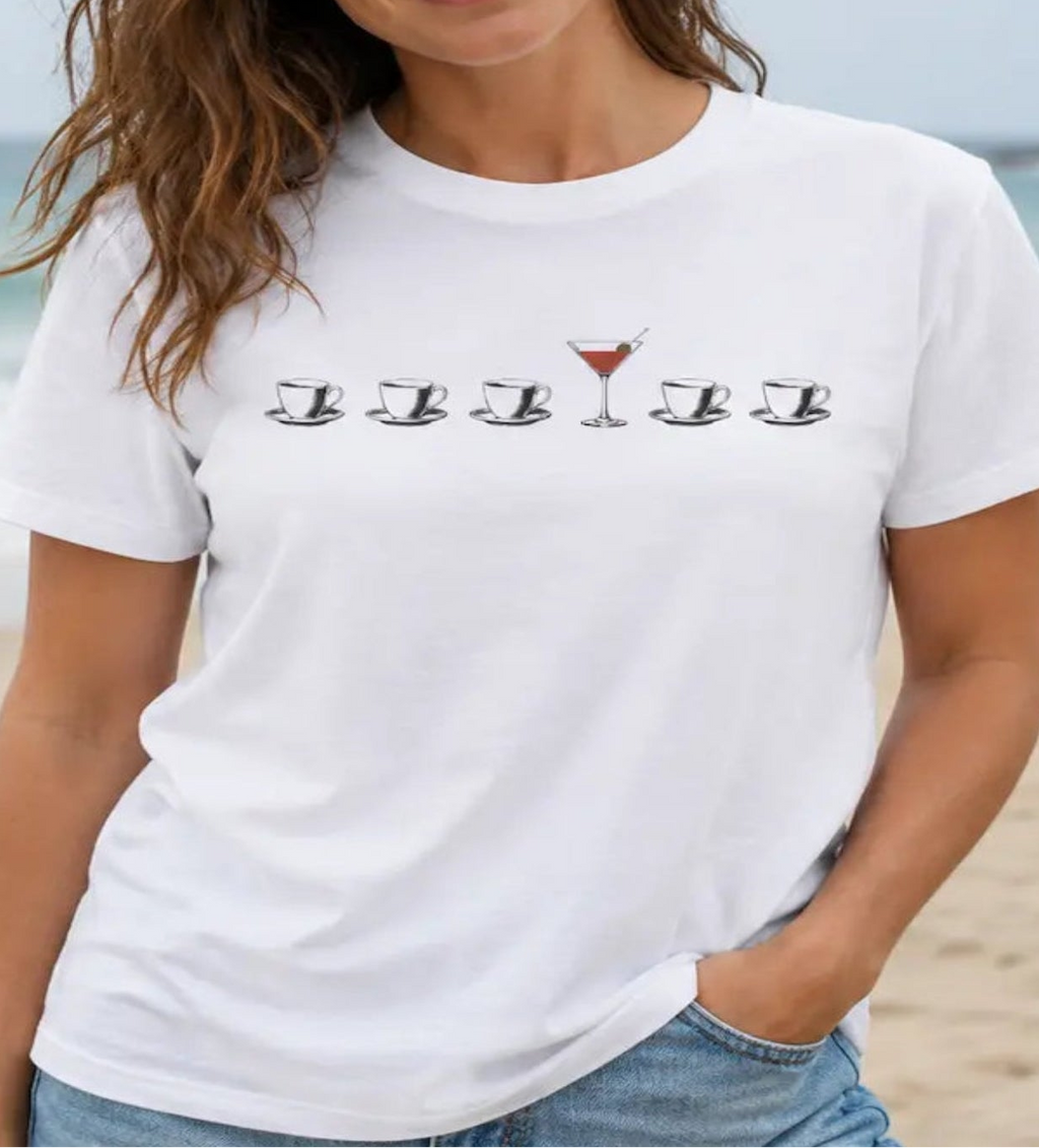 A woman wearing a white graphic t-shirt with a row of cocktail glasses and a martini glass design, standing on a sandy beach with the ocean in the background.