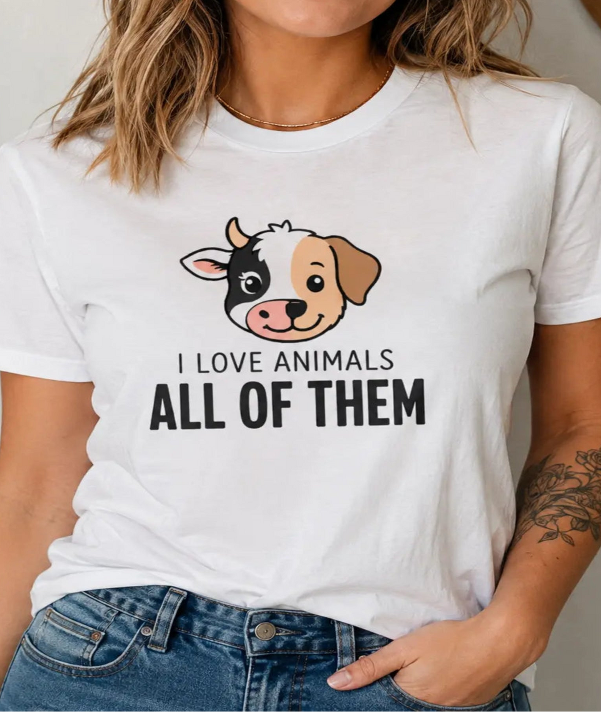 A woman wearing a white graphic t-shirt with a cartoon dog face and the text 'I LOVE ANIMALS ALL OF THEM' in black. She is also wearing blue jeans, standing in a brightly lit indoor setting.