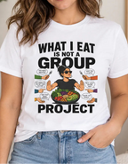 A woman wearing a white graphic t-shirt with a 'What I Eat Is Not A Group Project' design, paired with light-wash blue jeans in a cozy, well-lit indoor setting.