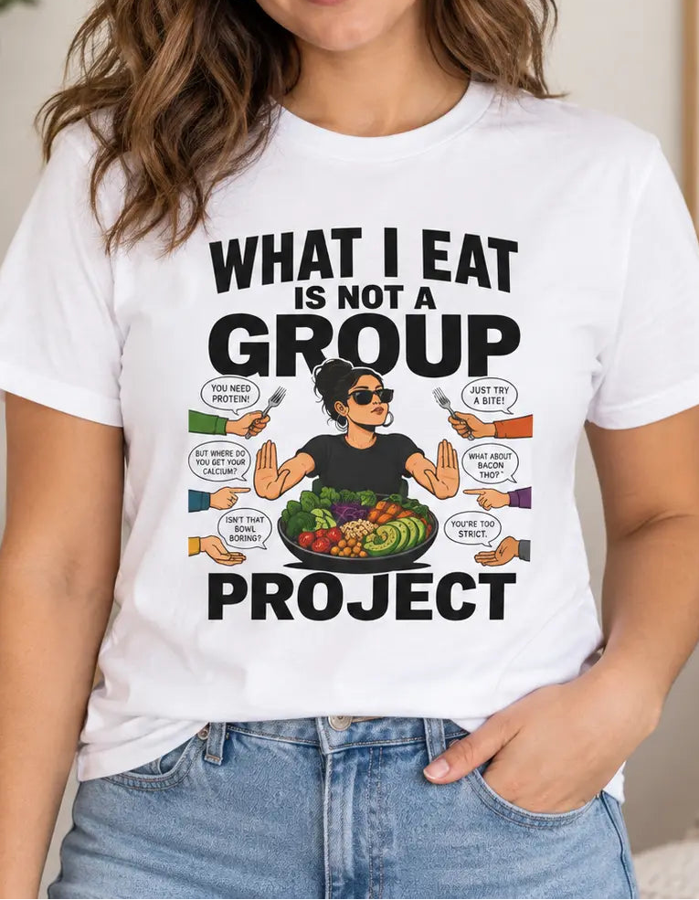 A woman wearing a white graphic t-shirt with a 'What I Eat Is Not A Group Project' design, paired with light-wash blue jeans in a cozy, well-lit indoor setting.