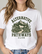 A woman wearing a white graphic t-shirt with a green mountain and forest design and the text 'ALTERNATIVE PATHWAYS CLUB'.