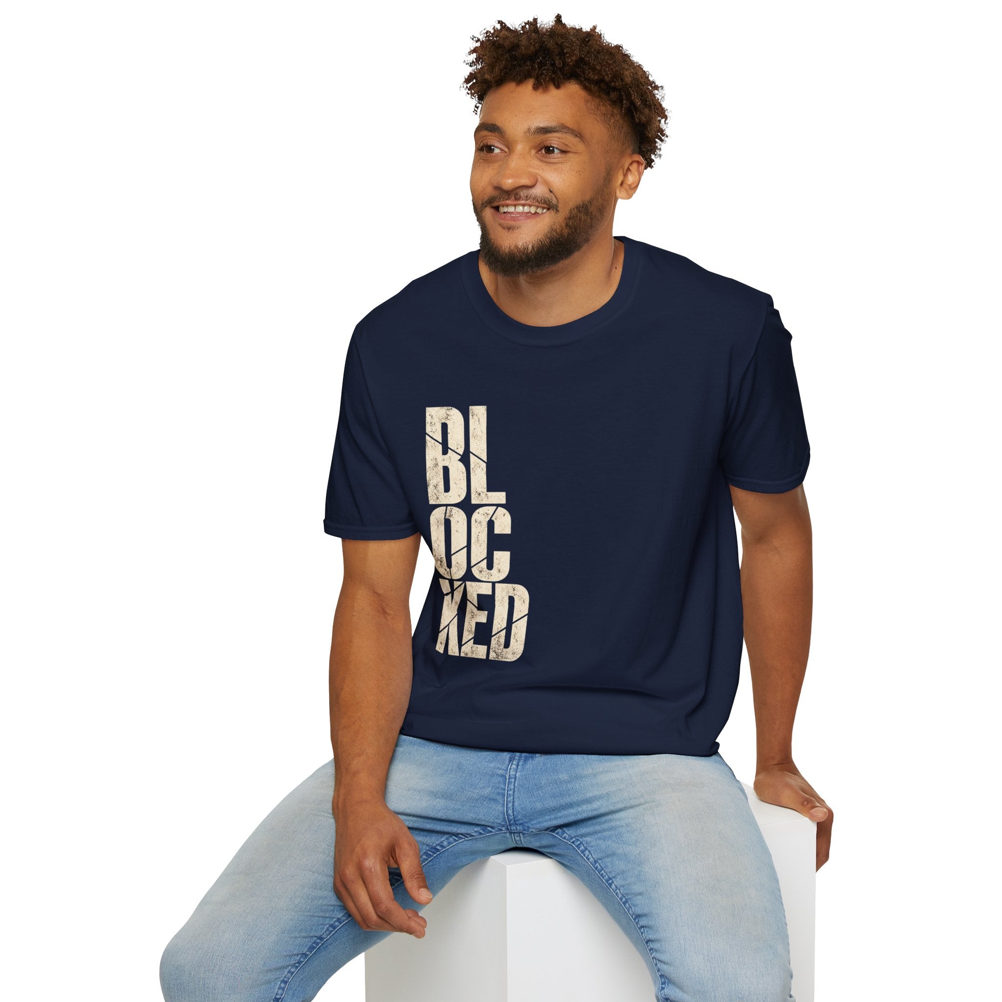 A man wearing a navy blue crewneck t-shirt with the word 'BLOCKED' printed vertically in distressed beige block letters, paired with light blue jeans, sitting on a white block against a white background.