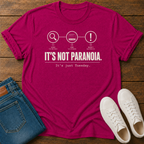 A hot pink short-sleeve graphic T-shirt laid flat on a wooden surface, styled with folded blue jeans and white sneakers.  text 'it's not paranoia it's a tuesday'
