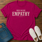 A bright magenta crewneck t-shirt with the words 'BECAUSE EMPATHY' printed in cream across the chest, styled in a flat lay on a dark wood background with blue jeans and white sneakers.
