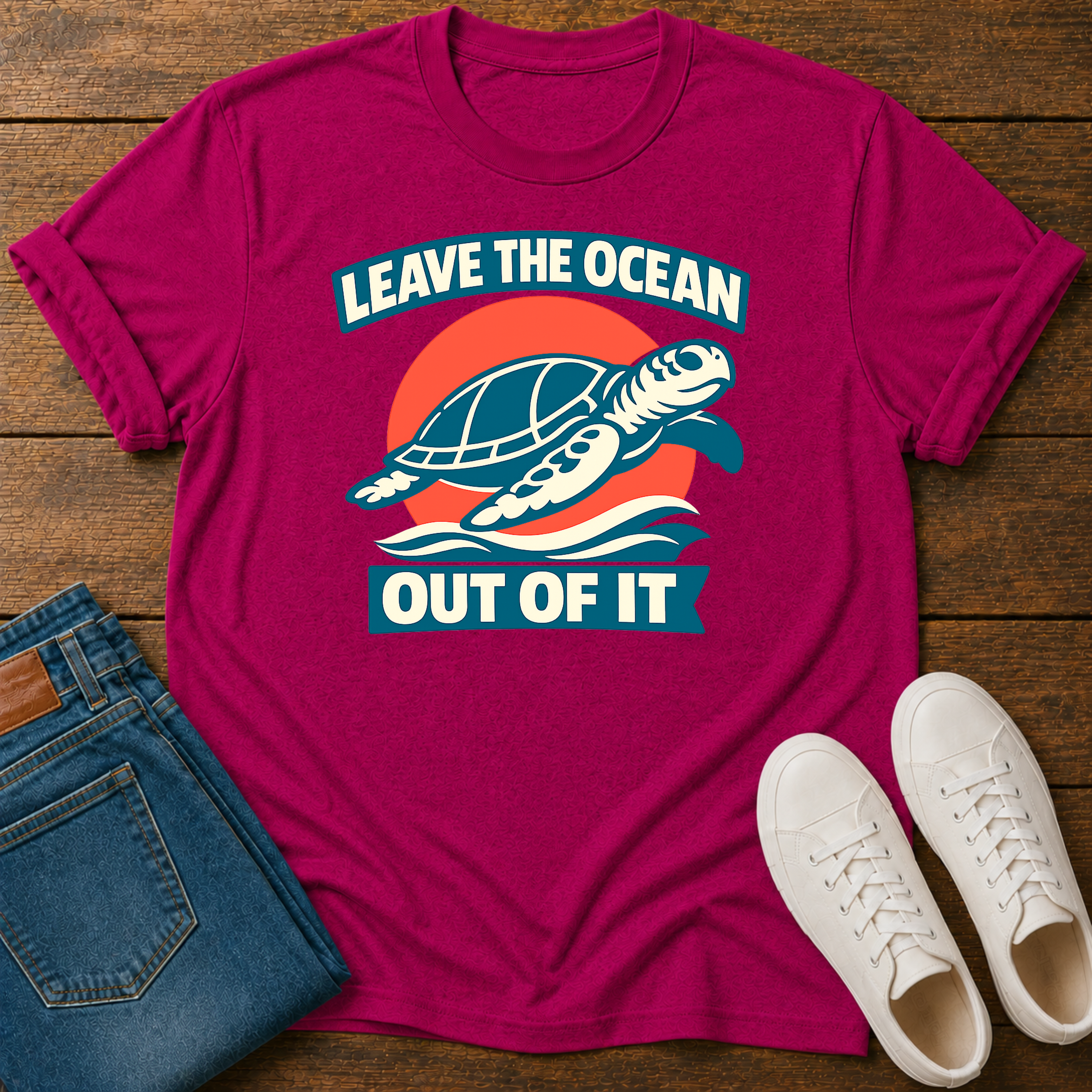 A magenta crewneck t-shirt with a retro turtle graphic and the text 'LEAVE THE OCEAN OUT OF IT', styled in a flat lay on a wooden background with blue jeans and white sneakers.