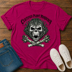 A bright pink graphic t-shirt laid flat on a wooden surface, styled with folded blue jeans and white sneakers. The t-shirt features a large green and black skull graphic with the text 'CERTIFIED PLANT MURDERER' arched above it.