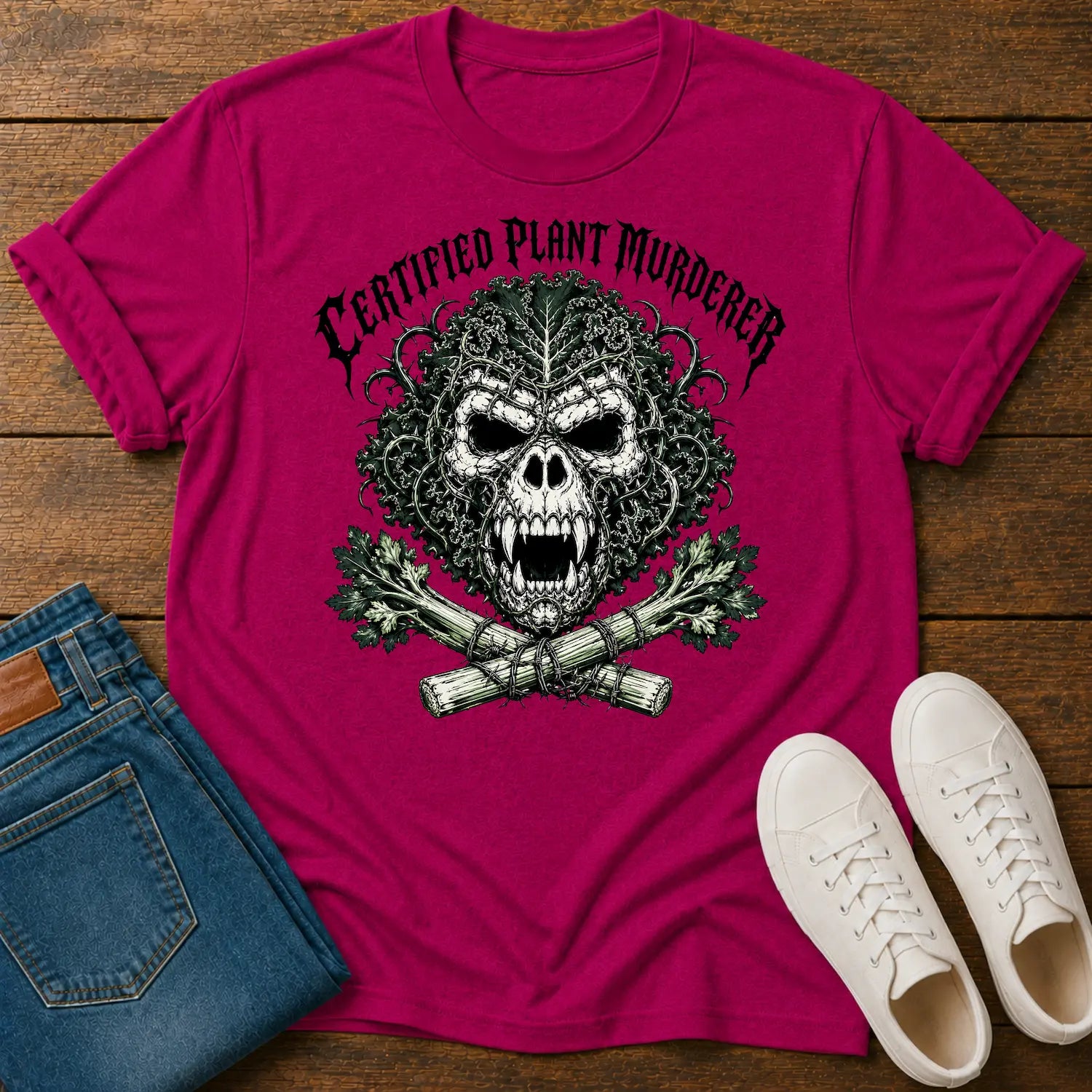A bright pink graphic t-shirt laid flat on a wooden surface, styled with folded blue jeans and white sneakers. The t-shirt features a large green and black skull graphic with the text 'CERTIFIED PLANT MURDERER' arched above it.
