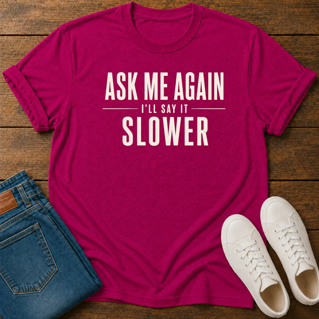 A bright pink graphic t-shirt with the text 'ASK ME AGAIN I'LL SAY IT SLOWER' in white, styled in a flat lay on a wooden background with blue jeans and white sneakers.