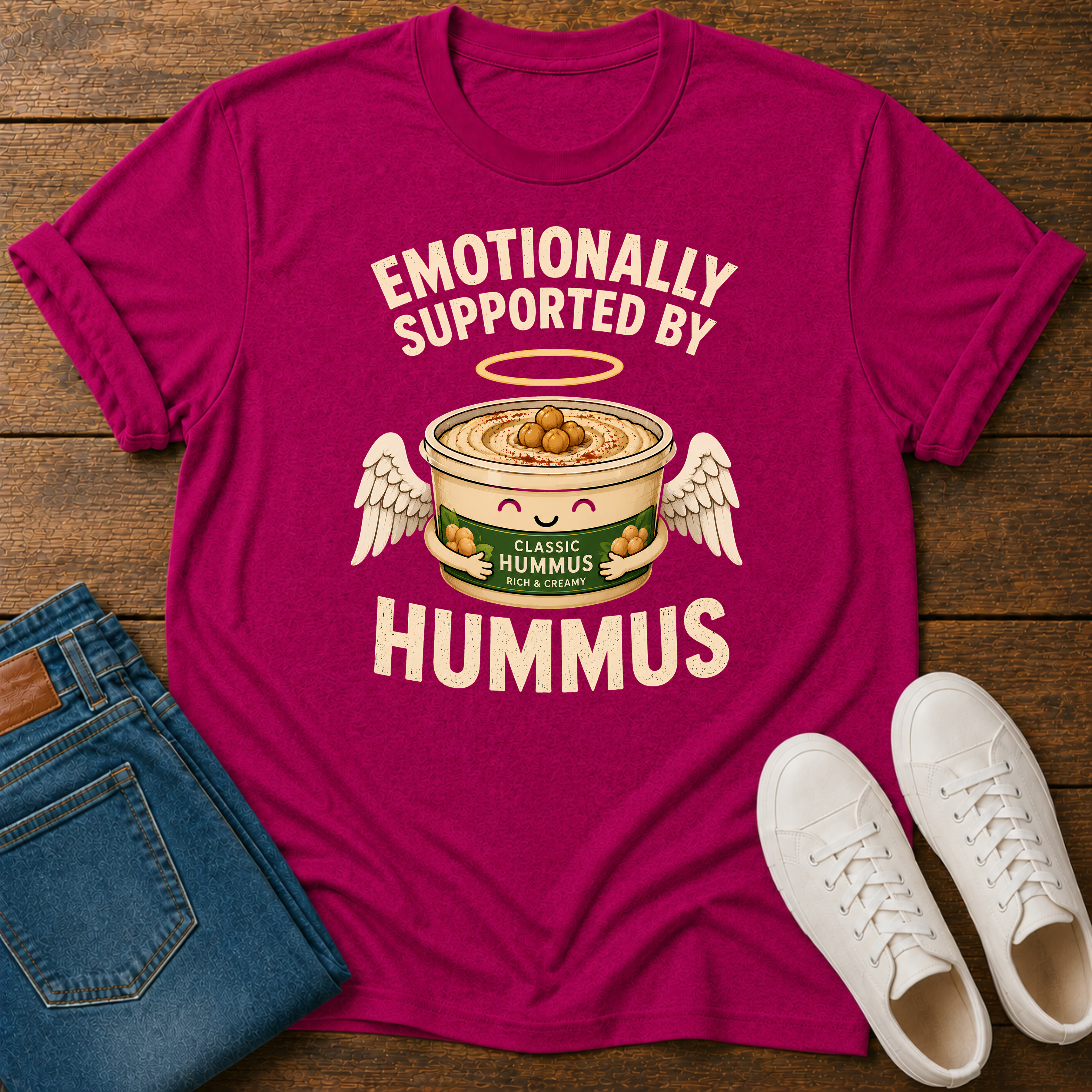 Hot pink graphic t-shirt with an image of a tub of hummus and text 'emotionally supported by hummus' on a wooden board with jeans and sneakers