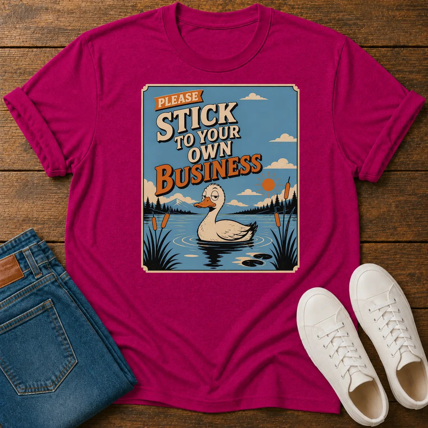 A bright magenta crewneck t-shirt with a retro-style duck graphic and the text 'PLEASE STICK TO YOUR OWN BUSINESS' laid flat on a wooden surface, styled with blue jeans and white sneakers.