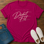 A hot pink graphic t-shirt laid flat on a wooden surface, styled with folded blue jeans and white sneakers. The t-shirt features the text 'MADE MYSELF SMALL ONCE. DIDN'T FIT'