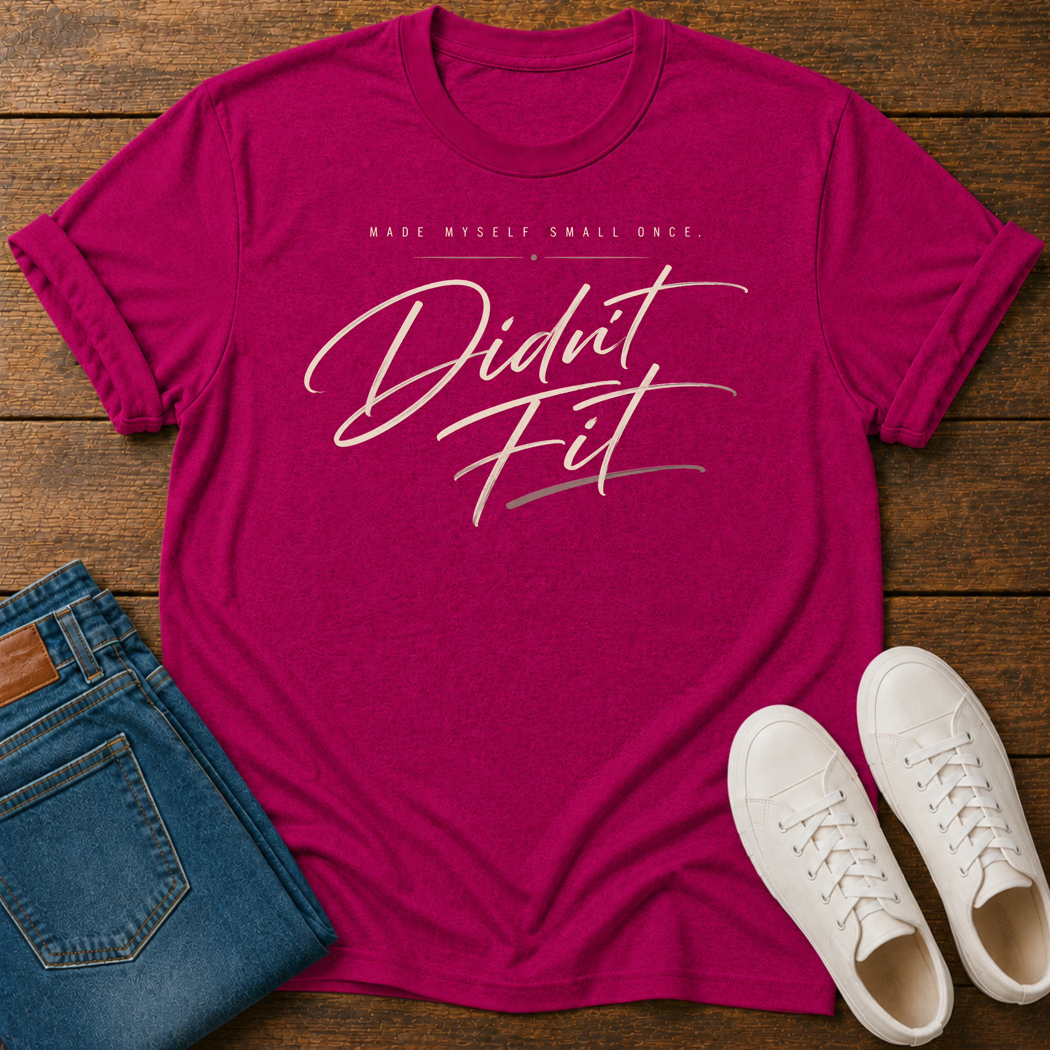 A hot pink graphic t-shirt laid flat on a wooden surface, styled with folded blue jeans and white sneakers. The t-shirt features the text 'MADE MYSELF SMALL ONCE. DIDN'T FIT'