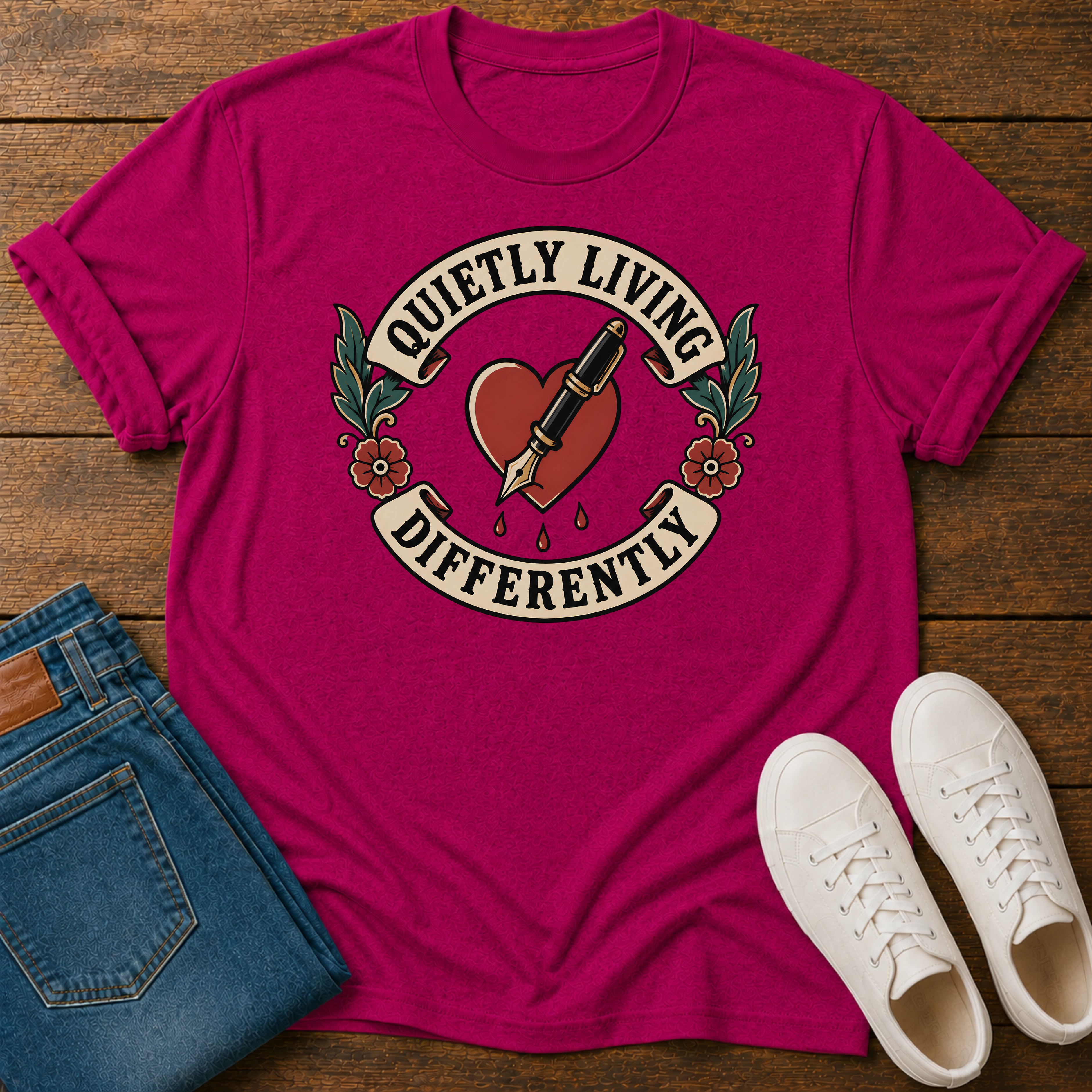 A bright pink graphic t-shirt laid flat on a rustic wooden background, styled with folded blue jeans and a pair of white sneakers. The t-shirt features a retro tattoo-style graphic of a red heart pierced by a black and gold fountain pen, with banners reading 'QUIETLY LIVING DIFFERENTLY' and green floral accents.