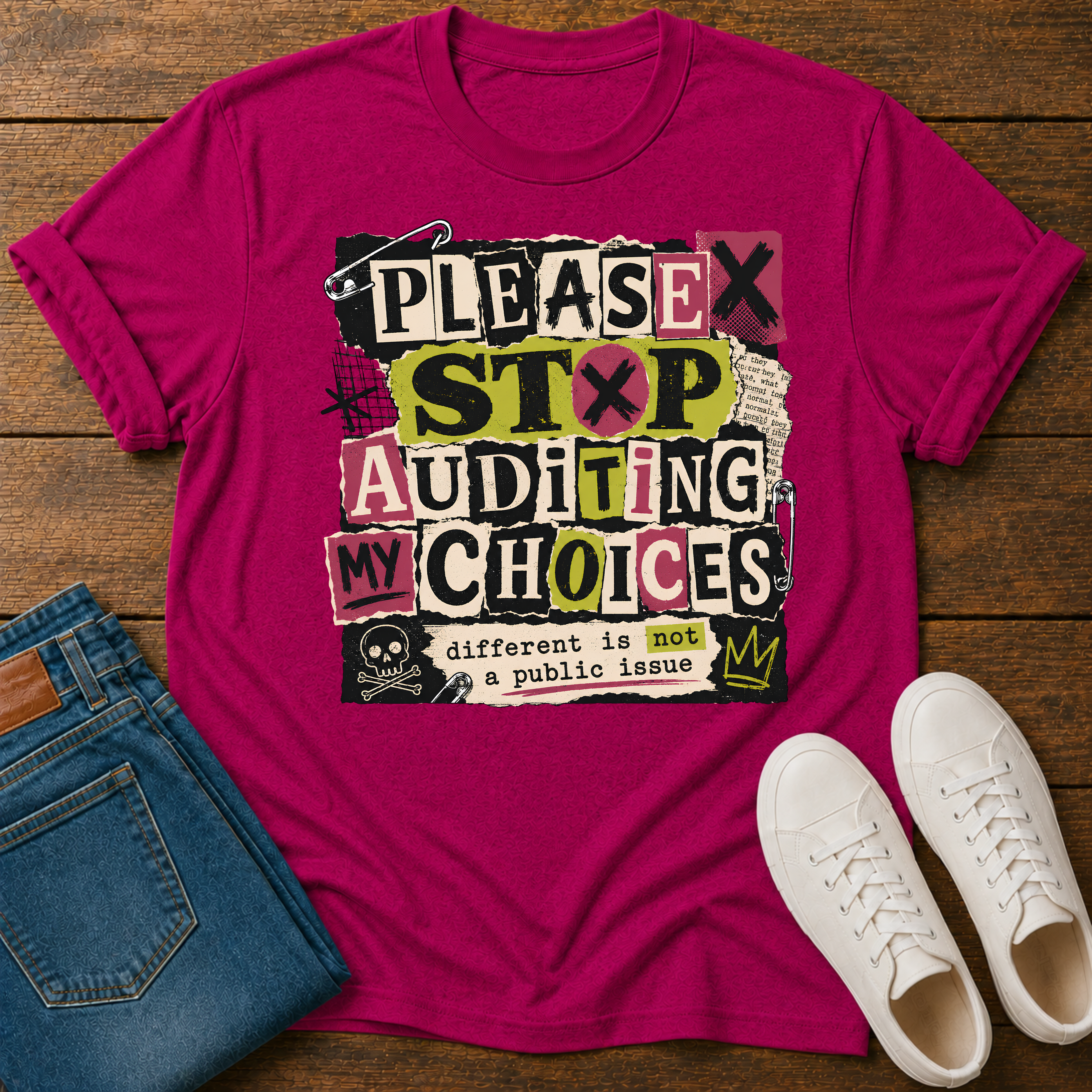 A bright pink graphic t-shirt laid flat on a wooden surface, styled with folded blue jeans and white sneakers. The t-shirt features a bold, multi-colored text graphic that reads 'PLEASE STOP AUDITING MY CHOICES' with a skull and crown detail.