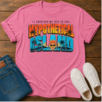 A pink short-sleeve graphic T-shirt laid flat on a wooden surface, styled with blue jeans and white sneakers. The shirt features a colorful retro-style text graphic reading 'I SURVIVED MY TRIP TO THE HYPOTHETICAL ISLAND' with palm trees and a cartoon face.
